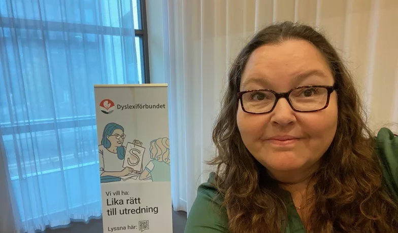 Kvinna med långt hår och glasögon. I bakgrunden en reklamvepa för Dyslexiförbundet.