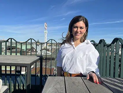 Kvinna (Rebecca Nobel) i vit skjorta på en takterass med blå himmel och NK-skylten bakom.